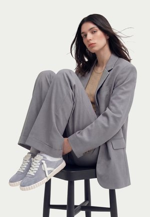 Young woman with long dark hair sits on black stool hugging knees, wearing gray suit, beige top, and light gray sneakers.