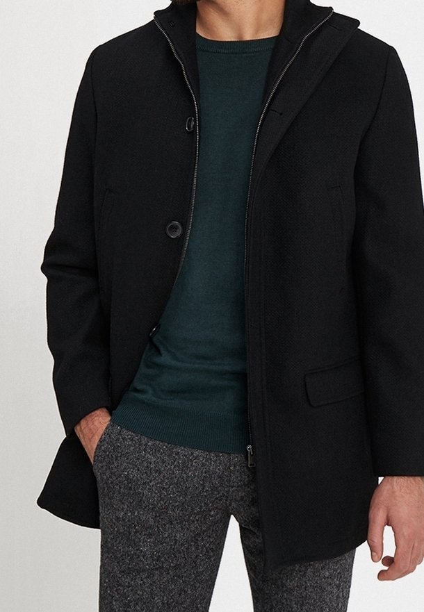 Černý kabát ze směsi vlny s límcem na stojáka, dvěma předními knoflíky a bočními kapsami. Nošen přes zelený svetr s kulatým výstřihem a šedé texturované kalhoty.