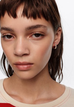 Joven con cabello corto y castaño, que lleva un pequeño aro de oro con piedras claras, maquillaje neutro y una blusa beige con detalles de costura en rojo.