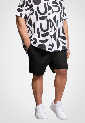 Mand iført sorte shorts, hvide sneakers og en hvid kortærmet skjorte med store sorte abstrakte former, stående med hænderne i lommerne.