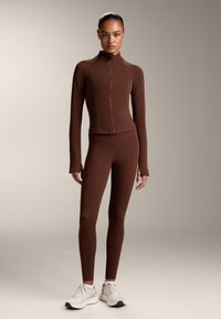 Smeđa atletska zip-up jakna i odgovarajuće tajice, izrađene od glatkog materijala. Bijeli tenisice s mrežastim detaljima upotpunjuju izgled.