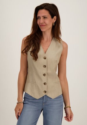 Vrouw met lang bruin haar die een beige gebuttoned vest en lichtblauwe jeans draagt, staand tegen een effen lichte achtergrond.