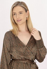 Rochie din material floral în verde închis, cu un model de flori maro deschis, decolteu în V, mâneci lungi cu manșete elastice și talie legată.