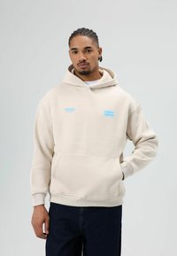 Beige hoodie met een voorzak en geribbelde boorden. Heeft blauwe logo's "STRONGER" en "chance" op de borst. Zachte stoftextuur.