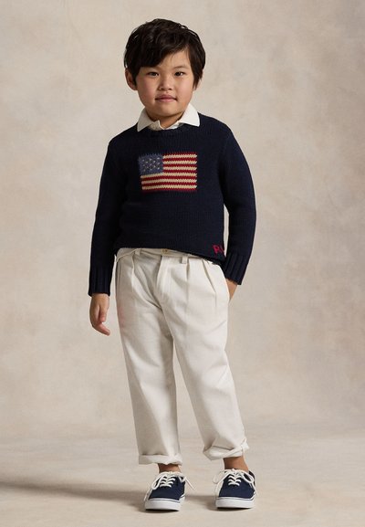 Young child standing wearing navy sweater with an American flag, white collared shirt, cream pants, and navy sneakers against a plain background.