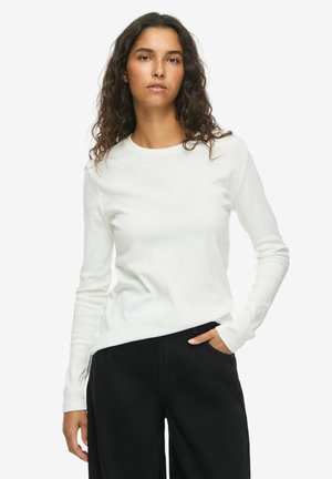Femme aux cheveux bouclés portant une chemise blanche à manches longues et un pantalon noir, debout avec une main dans la poche sur un fond uni.