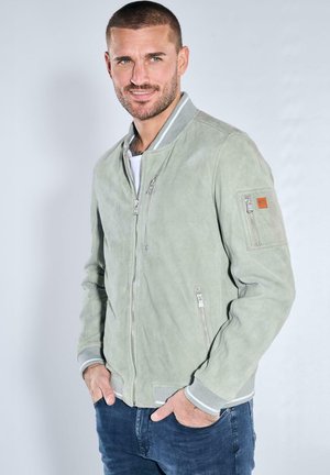 Mann mit kurzen Haaren und Bart, der eine hellgraue Bomberjacke mit Reißverschluss und blaue Jeans trägt, lächelt mit den Händen in den Taschen.