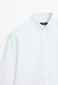 Weißes Hemd mit Knopfleiste, klassischem Kragen und langen Ärmeln. Hergestellt aus glattem Stoff mit feiner, dezenter Textur. Die Knöpfe sind transparent.