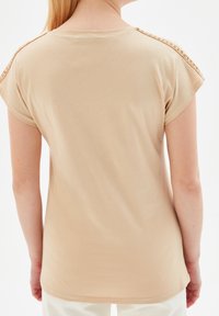 Beige top s krátkým rukávem a kulatým výstřihem; má dekorativní zapletené detaily na ramenech. Vyrobeno z měkké, lehké tkaniny.