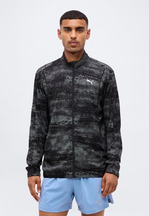 Mann trägt eine schwarz-grau gemusterte Jacke mit Reißverschluss, weißem Puma-Logo und hellblauer Puma-Shorts und steht vor einem schlichten Hintergrund.