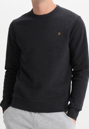 Hombre con un suéter de manga larga gris oscuro con un pequeño logo naranja "F" en el pecho y pantalones deportivos gris claro, manos en los bolsillos.