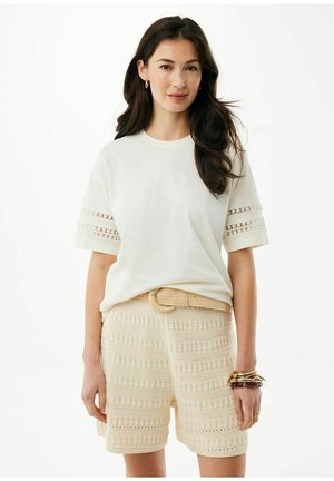 Jonge vrouw met lang donker haar draagt een witte top met korte mouwen en kantdetails, crèmekleurige gehaakte korte broek met riem, en meerdere armbanden aan haar rechterpols.