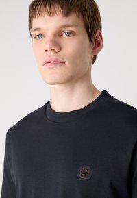 Černé bavlněné tričko s kulatým výstřihem, na levém prsu má kulatou nášivku s logem. Hladká textura a přiléhavý střih.