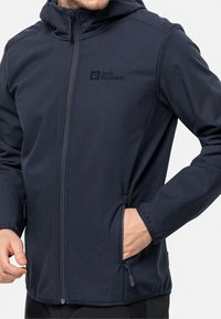 Veste softshell bleu marine avec fermeture éclair sur le devant, capuche, poches latérales et un logo sur la poitrine gauche. Elle présente une texture lisse.