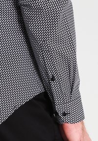 Mustavalkokuosinen pitkähihainen paita, jossa on geometrinen kuvio, nappikiinnitteiset kalvosimet ja sileä pinta.