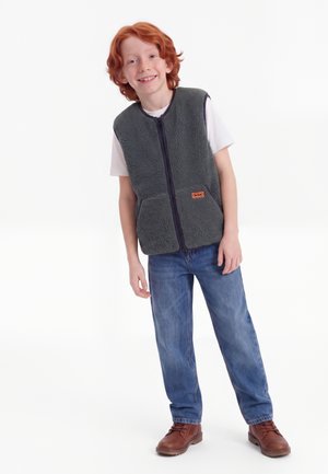 Rødhåret barn smiler, iført en grå fleecevest med lynlås, hvid t-shirt, blå jeans og brune støvler, stående mod en hvid baggrund.