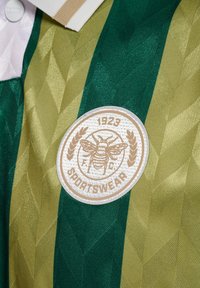Grøn og guld sportstrøje med en geometrisk tekstur, der har et cirkulært broderet mærke med en bi og "1923 SPORTSWEAR."