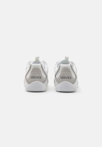 Zapatillas de cuero blancas con acentos de ante gris, que presentan una suela texturizada y una lengüeta en el talón. Nombre de la marca "Versace" impreso en gris en la parte posterior.