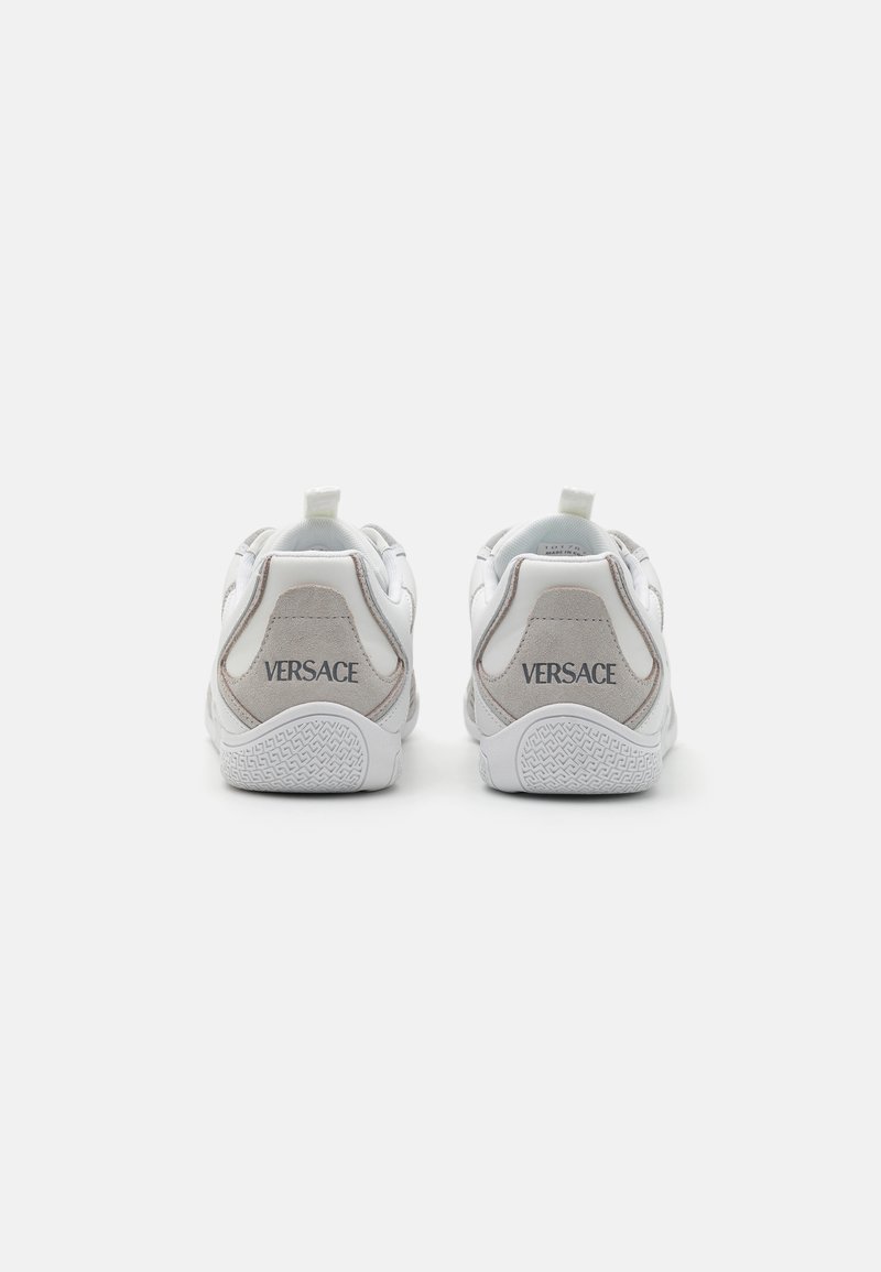 White leather sneakers with gray suede accents, featuring a textured sole and a heel tab. Brand name "Versace" printed in gray on the back.