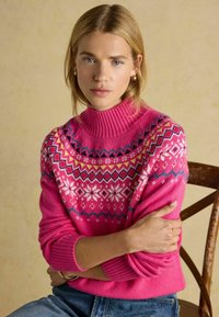 Fuksijasti pletena pulover s visokim rebrastim ovratnikom, ki ima barvite geometrijske in cvetlične vzorce na sprednjem delu. Mehka tekstura in sproščen kroj.