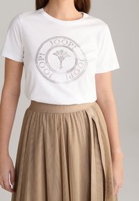 Camiseta blanca de algodón con un logo redondo y texturizado que presenta un diseño floral. Combinada con una falda beige con pliegues y una cinturilla con marca.
