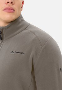 Giacca in fleece grigia con colletto alto, chiusura con zip e logo nero sul petto. Texture liscia e design aderente.