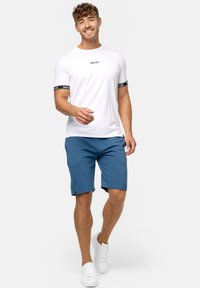 Witte t-shirt met zwarte strepen en logo op de mouwen, gecombineerd met blauwe shorts, gedragen met witte sneakers. Casual, sportieve outfit.