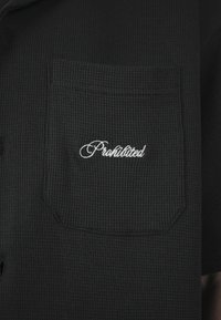 Tissu noir texturé avec une poche poitrine brodée d'un texte en écriture cursive blanche indiquant "Interdit."