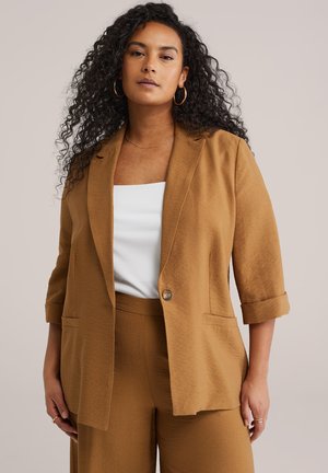 Vrouw met krullend haar die gouden oorringen draagt, een zandkleurig colbert met opgerolde mouwen, bijpassende broek en een wit geribd topje.