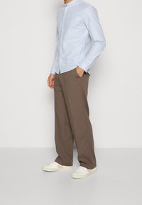 Camisa de botón de manga larga azul claro con collar alto, combinada con pantalones de pierna ancha marrones y zapatillas blancas con detalles claros.
