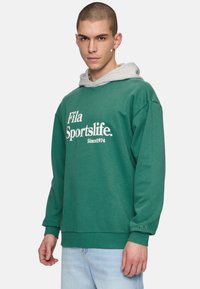 Felpa verde con cappuccio grigio, con la scritta "Fila Sportslife" in bianco. Maniche e orlo a costine, texture morbida, vestibilità rilassata.