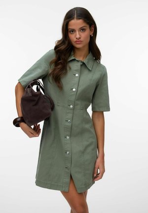 Ženska v olivno zeleni obleki s kratkimi rokavi z gumbi, ki drži temno rjavo torbico, z valovitimi dolgimi lasmi in nevtralnim izrazom obraza.