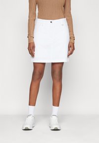 Persoon met een witte knielange rok, beige geribd langarmshirt, witte sokken en witte sneakers, staand tegen een effen achtergrond.