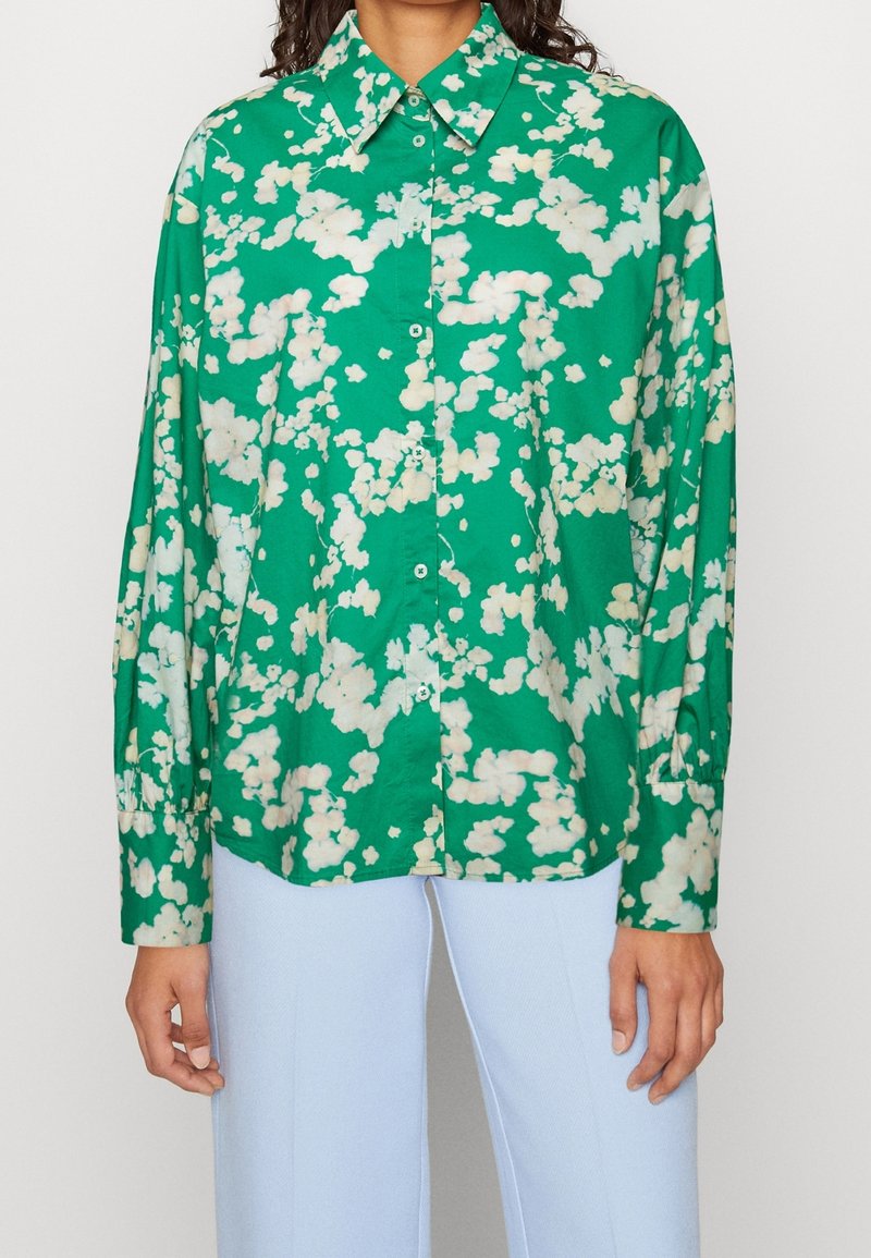 Chemise verte à manches longues boutonnée avec un motif abstrait blanc évoquant des nuages, portée avec un pantalon bleu clair.