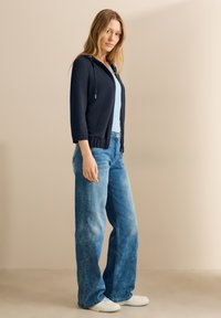 Felpa navy con cappuccio e maniche a tre quarti, abbinata a una maglietta azzurro chiaro e a jeans a gamba larga blu sbiaditi, su uno sfondo neutro.