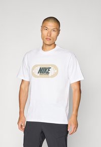Biała bawełniana koszulka z okrągłym dekoltem, z centralnym nadrukiem sztangi do podnoszenia ciężarów z napisem "NIKE" w kolorze zielonym i beżowym.