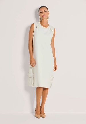 Frau trägt ärmelloses cremefarbenes Midikleid mit Schulterverzierungen und seitlichen Klappentaschen, kombiniert mit beigen spitzen Absätzen und großen Ohrringen.