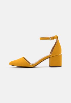 Sandalias de ante amarillas con tacón cuadrado, punta afilada y correa en el tobillo, que presentan un diseño elegante y una textura suave.