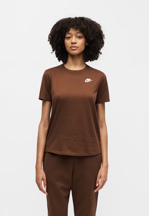 Ung kvinde med krøllet hår iført en brun Nike T-shirt og matchende brune bukser, står foran en ensfarvet lys baggrund.