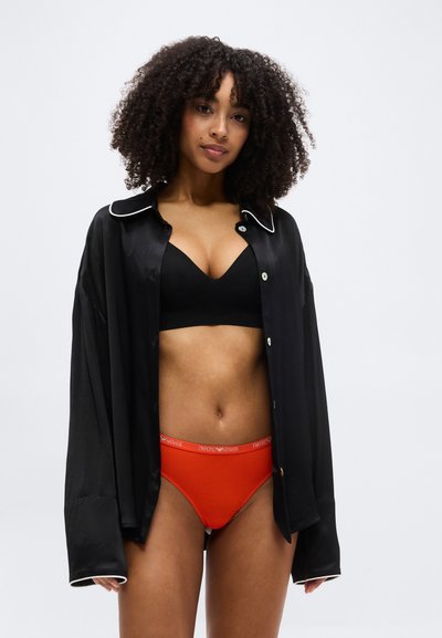 Femme aux cheveux bouclés portant un soutien-gorge noir, un sous-vêtement rouge avec une ceinture à logo, et une chemise noire à manches longues ouverte, sur un fond uni.