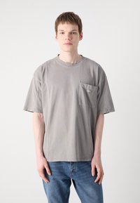 Grå bomulds T-shirt med kortærmer, der har en brystlomme, rund halsudskæring og løs pasform, vist sammen med blå denimjeans.