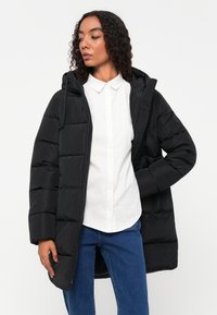 Schwarze Pufferjacke mit Kapuze, ausgestattet mit einem gesteppten Design, Reißverschluss, Seitentaschen und langen Ärmeln. Trägt man über einem weißen Hemd mit Kragen.