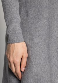 Tessuto a maglia grigio a coste con una manica aderente, in primo piano una mano che riposa contro il tessuto; texture liscia e aspetto morbido.
