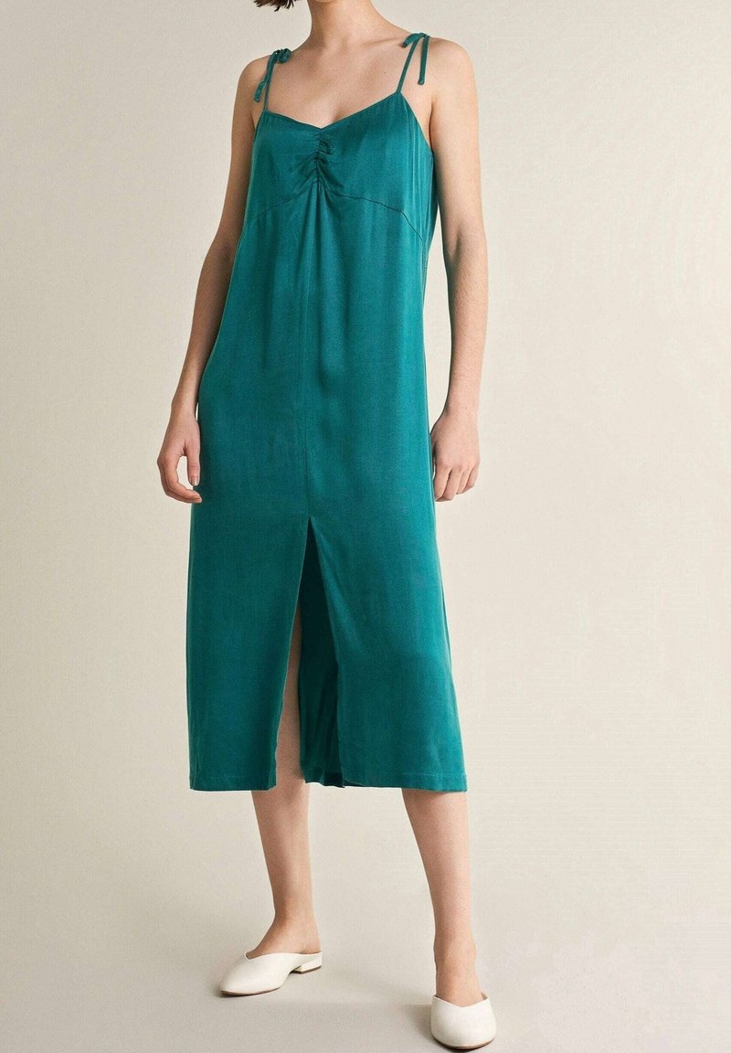 Robe midi sans manches couleur sarcelle avec bretelles réglables, détail froncé à l'avant et fente frontale. Le modèle la porte avec des mules blanches.