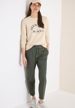 Žena opírající se o bílou zeď, má na sobě béžovou čepici, béžovou mikinu s nápisem "Seek Places", olivově zelené kalhoty se stahovací šňůrkou a stříbrné tenisky.