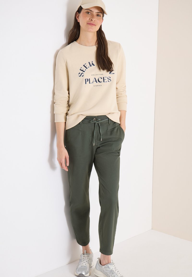 Kvinde lænende sig op ad en hvid væg iført beige kasket, beige sweatshirt med teksten "Seek Places," olivengrønne bukser med snøre og sølvfarvede sneakers.