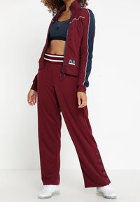 Bordowy sportowy dres z dopasowaną kurtką i spodniami o szerokich nogawkach. Kurtka ma granatowe akcenty i zamek błyskawiczny. Spodnie wyposażone są w zatrzaski po bokach.