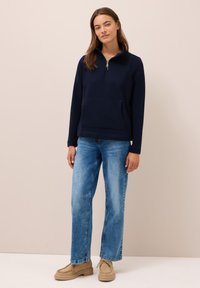 Donna in piedi con un pullover navy a zip fino al collo, jeans dritti azzurri chiari e scarpe beige con lacci, contro uno sfondo beige semplice.