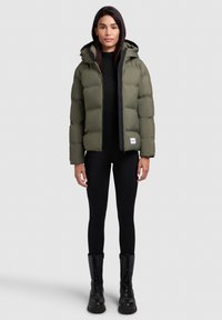 Puffjakke i olivengrøn med hætte, lynlås lukning og sorte detaljer. Bæres over en sort top og tights, suppleret med sorte snørebåndsstøvler.