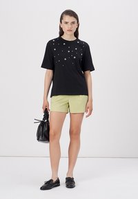 Sort T-shirt med sølvstjerner, korte ærmer, parret med lysegrønne shorts. Modellen holder en sort taske og har sorte sko på.